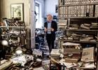 Der Historiker und Professor steht mit einem Druckwerk zwischen zwei Räumen, die weitgehend mit Studienmaterialien vollgeräumt sind.