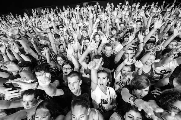 Weitwinkelbild von einer Menschenmasse - Junge Menschen die jubelnd die Hände in die Höhe strecken. in der Bildmitte blickt eine junge Frau in die Kamera, streckt die Zunge raus und zeigt Daumen hoch. Schwarz-Weiß.