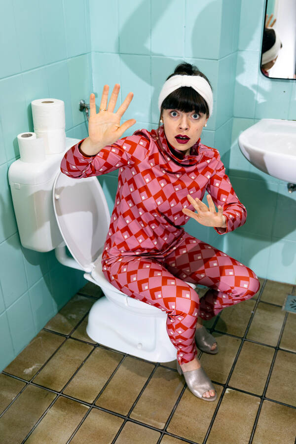 Eine Frau in rot gemustertem Einteiler sitzt auf einer Toillette und blickt erschrocken in die Kamera, die Hände abwehrend erhoben. Hellblaue Wand und braune Bodenfliesen.