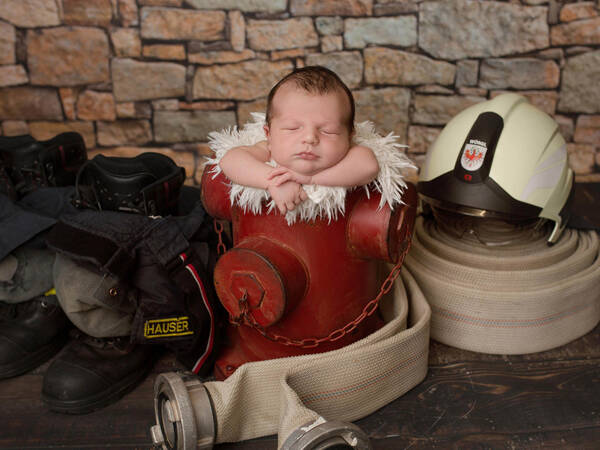 Ein Baby liegt auf einem roten Hydranten, links und rechts davon sind Feuerwehr-Ausrüstung aufgelegt.