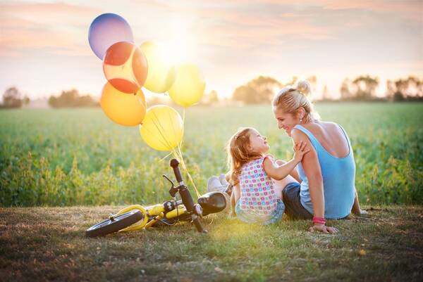Eine Frau und ein Mädchen sitzen neben einem Rad und Luftballons in der Abendsonne an einem Weg am Boden.