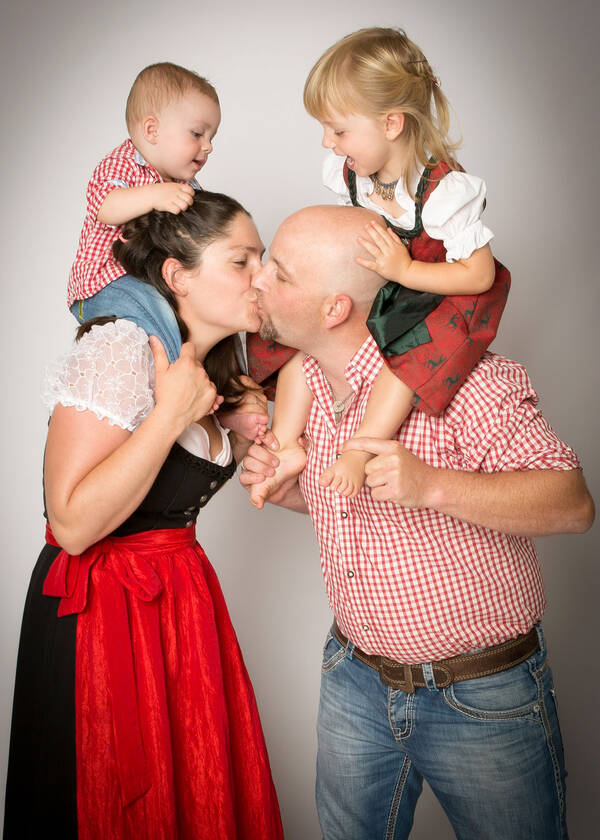 Ein Mann und eine Frau küssen sich. Der Mann hat ein Mädchen auf den Schultern und die Frau hat einen kleinen Buben auf der Schulter. Die Frau und das Mädchen tragen Trachtenkleider, der Mann und der Bub tragen Jeans und weißrot karierte Hemden.