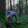 Eine am ganzen Körper tätowierte Person sitzt nackt mit dem Rücken zum Fotografen auf der moosigen Wurzel eines Baumes im Wald.