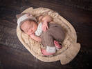 ein neugeborenes Baby liegt schlafend in einem kleinen Holztrog. Es trägt braune, spitzenbesetzte Strickwäsche und -haube.
