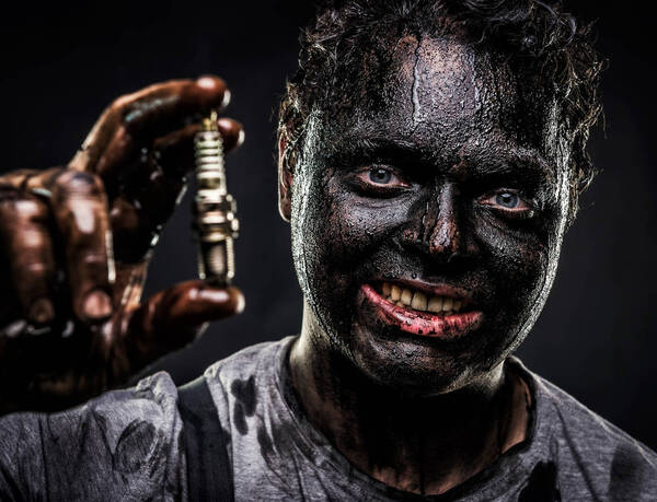 Ein Mann mit völlig schwarz verschmiertem Gesicht hält lächelnd eine Zündkerze in der Hand.