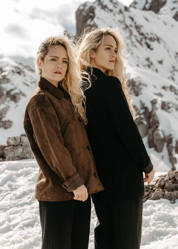 Zwei blonde Frauen stehen in schneebedeckter Berglandschaft.