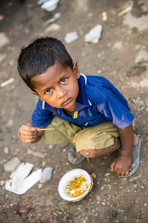 Ein dunkelhäutiges Kind kauert am Boden und schaut nach oben in die Kamera. Auf der Erde vor ihm steht eine Schüssel mit etwas Essen.