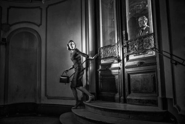 Eine Frau geht über eine Treppe und sieht sich besorgt um. Durch eine Glastüre ist ein Mann zu sehen. Schwarz-Weiß.