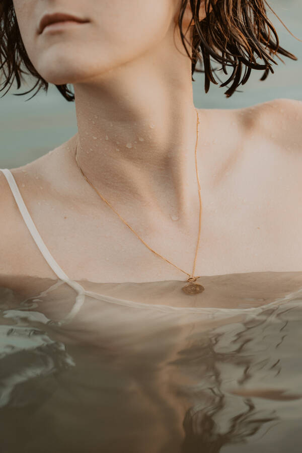CloseUp einer Frau die mit einem Trägerleibchen im Wasser steht. um den Hals hat sie eine rotgoldene Kette mit einem Anhänger der schon unter die Wasseroberfläche hängt.