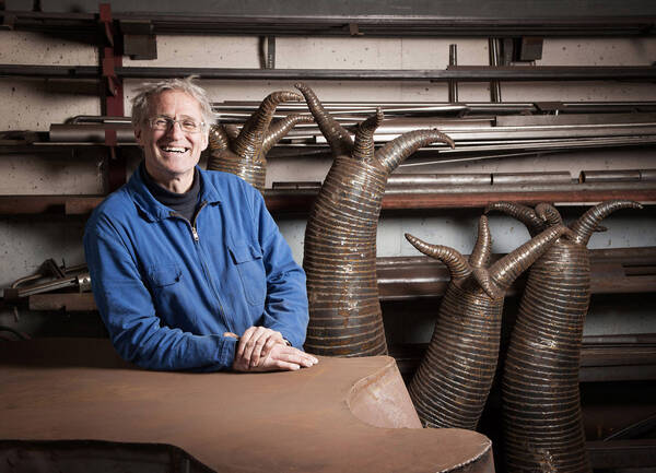 Ein Mann lehnt lachend im blauen Arbeitsanzug auf einer Metallskulptur. Mehrere Tentakeltragende Skulpturen stehen hinter ihm vor einem Regal mit unterschiedlichen lange Metallteilen. 