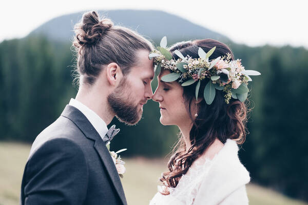 ein Brautpaar steht sich gegenüber. Sie mit einem Blumenkranz, er mit langen, aufgesteckten Haaren und Bart. Ihre Nasenspitzen berühren sich.