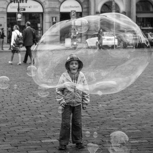 Ein Kind mit Kapuzenjacke auf einer gepflasterten Straße schaut voller Begeisterung durch eine enorme Seifenblase. Schwarz-Weiß.
