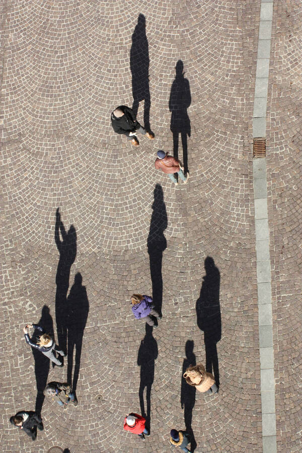 Blick von oben auf Menschen auf einem gepflasterten Platz. Die tiefstehende Sonne zeichnet lange Schatten der Menschen auf das Pflaster.