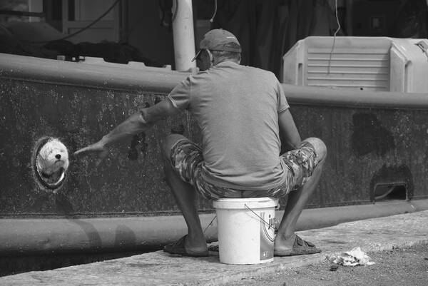 Ein Mann sitht auf einem Plastikkübel an einer Hafenmole. aus einem Bullauge des danebenliegenden Schiffes schaut ein Hund heraus. Schwarz-Weiß.