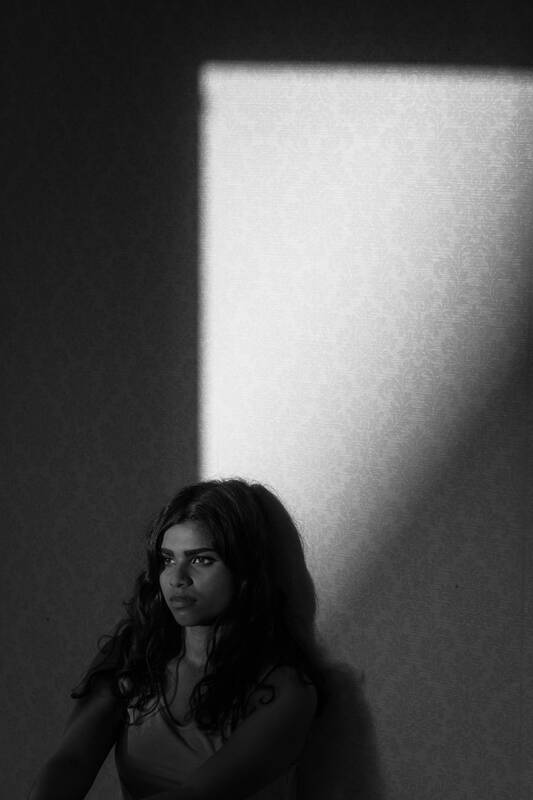 Junge Frau an eine ornamental gemusterte Wand gelehnt, Licht fällt durch ein Fenster auf ihr Gesicht. Schwarz-Weiß