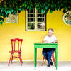Eine ältere Frau sitzt vor einer gelben Hauswand mit Fenster auf einem roten Sessel an einem grünen Tisch und liest.