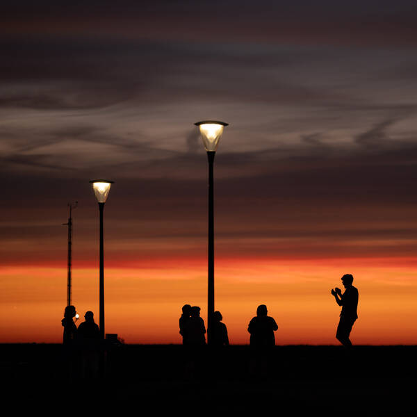 Silhouetten mehrerer Menschen im Abendlicht unter bereits leuchtenden Straßenlaternen.