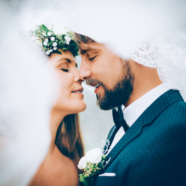 Braut mit weiß-grüner Blumenkrone und bärtiger Bräutigam kurz vor einem Kuss unter dem Schleier