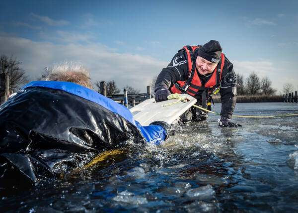 Ein Rettungseinsatz auf dem Eis: Ein Helfer mit einem Schwimmboard kniet auf dem eis. im Vordergrund versucht sich ein Mann, der im Wasser ist, an dem Board festzuhalten.