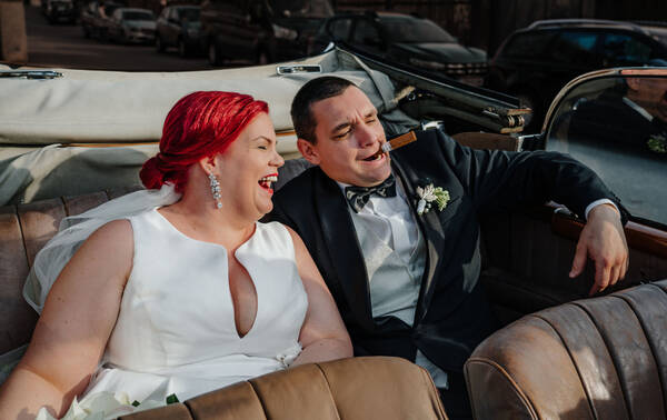 Bild eines Brautpaares in einem Oldtimer Cabrio. Der Mann hat eine Zigarre im Mund. Die Braut hat knallrote Haare und lacht. 