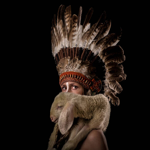 Eine Frau mit indianischem Federschmuck und weißer Bemalung im Gesicht hat ein Lamm über die Schultern gelegt.
