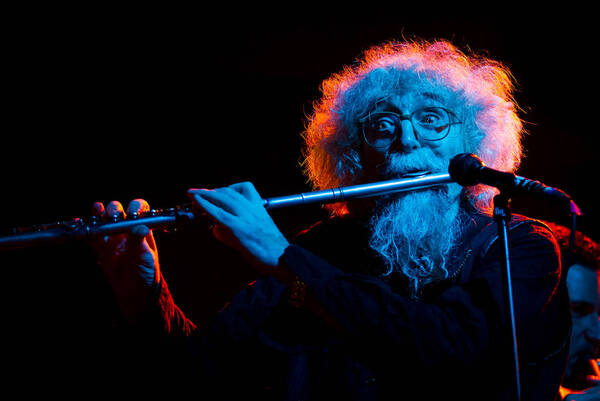 Weißbärtiger Musiker mit Querflöte bei einem Livekonzert  vor dem Mikrofon. Von hinten rot beleuchtet, von vorne blau
