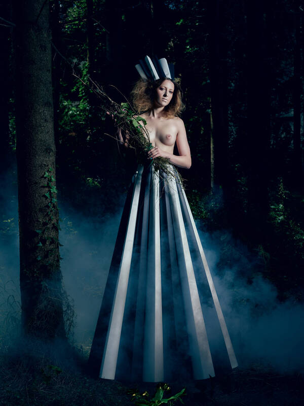 Teilakt – eine Frau steht mit einem Strauß belaubter Zweige in einem nebligen Wald. Der Oberkörper ist unbekleidet, am Kopf hat sie einen schwarz-weiß-gestreiften Hut und der Unterleib ist in einem überdimensional langen schwarz-weiß-gestreiften Rock.