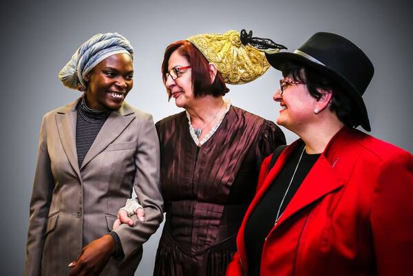Drei ältere Frauen, eine mit dunkler Hautfarbe und afrikanischer Kopfbedeckung, eine mit Oberösterreichischer Goldhaube und eine mit schwarzem Hut.