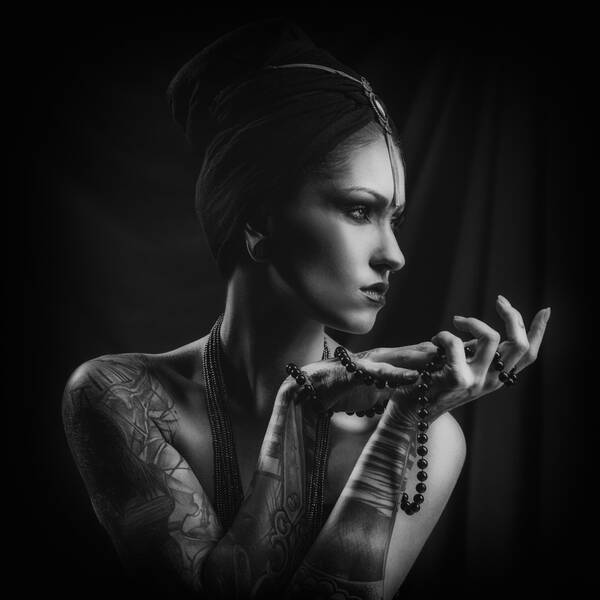 eine tätowierte Frau mit vielen Ketten deren Haare mit einem Tuch nach oben gebunden sind, schaut nach rechts aus dem Bild. Schwarz-Weiß.