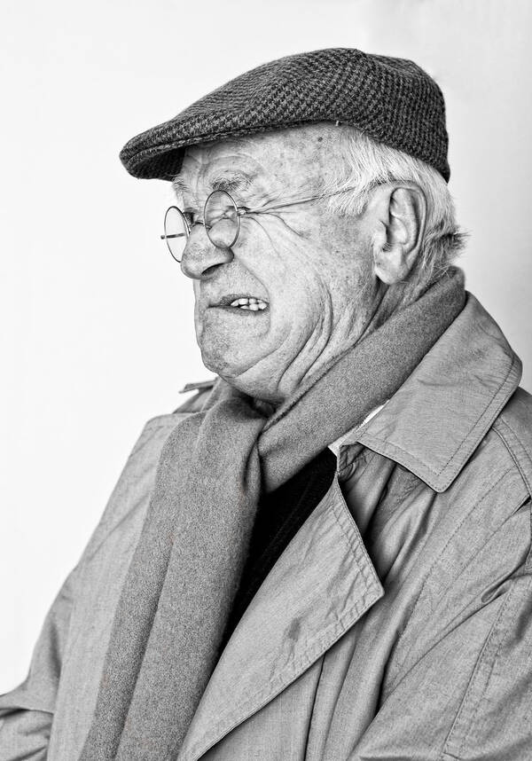 Ein alter Mann im Trenchcoat mit Schal, Nickelbrille und Schirmmütze verzieht kritisch das Gesicht. Schwarz-Weiß.
