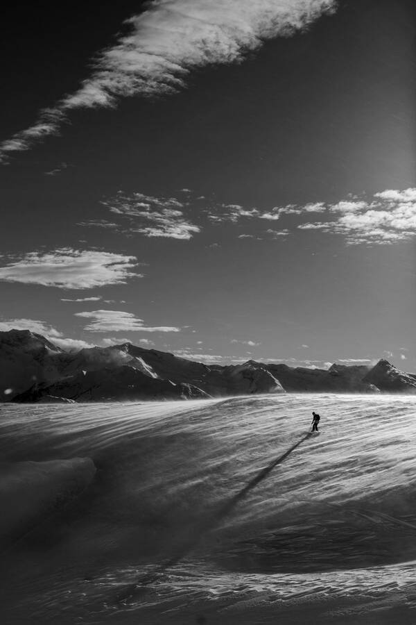 Jemand geht im winterlichen Gebirge bei starken Schneeverwehungen über einen Geländerücken. Die tiefstehende Sonne wirft einen langen Schatten der Person über den ganzen Hang. Schwarz-Weiß.