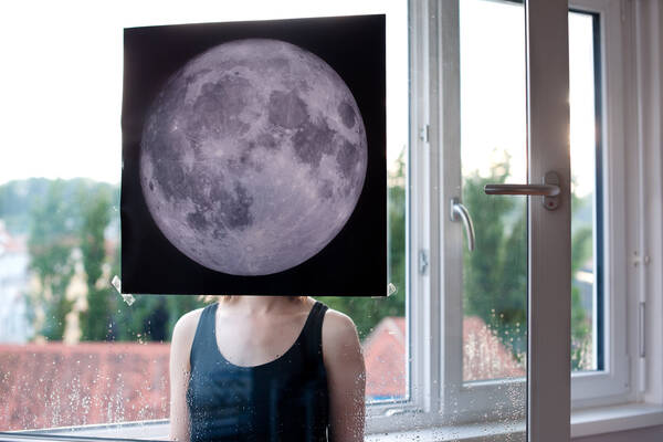 Eine Frau steht vor einem Fenster. Auf dem Fenster klebt ein Foto vom Mond und verdeckt das Gesicht der Frau.