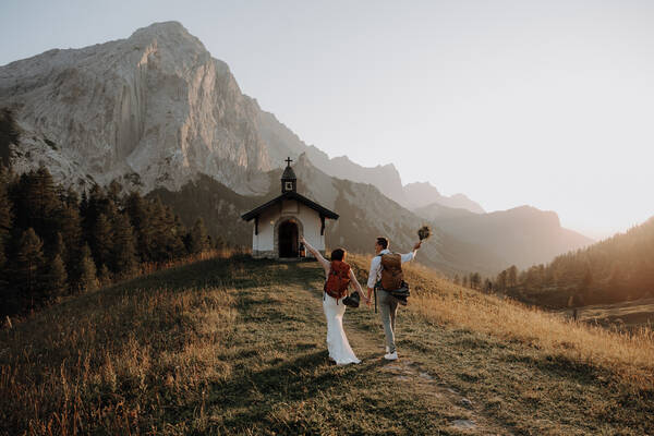 Eine Braut in weißem Kleid und der Bräutigam wandern mit roten Rucksäcken zu einer Bergkapelle. 