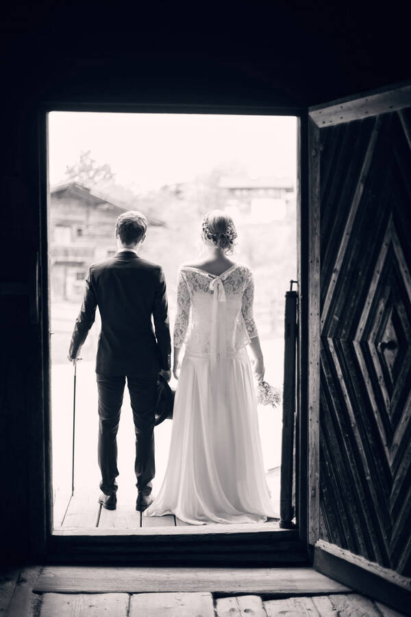 Ein Brautpaar steht vor der Tür eines alten, rustikalen Hauses. Blick von innen. Schwarz-Weiß.