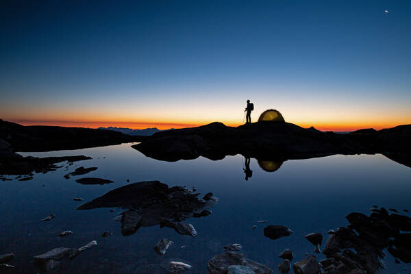 Silhouette eines Wanderers neben seinem Zelt vor dem Abendhimmel, der sich im Wasser des Vordergrundes spiegelt.