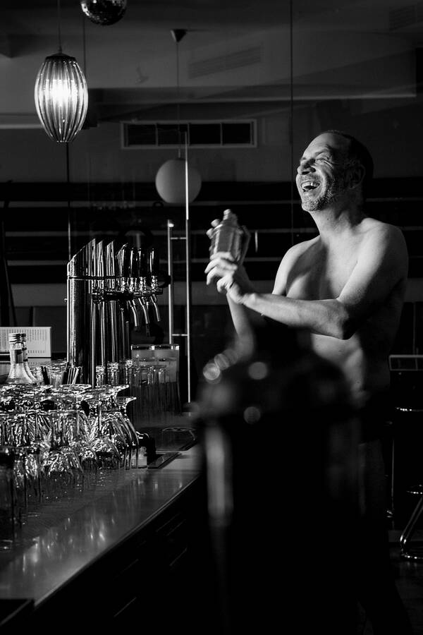 Ein nackter Mann steht hinter einer Bar und mixt einen Cocktail. Schwarz-Weiß.