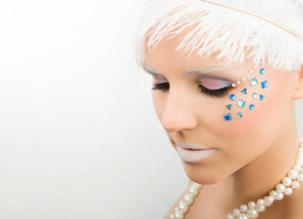 High-Key-Portrait einer ungen Frau mit weißer gefiederter Kopfbedeckung, weiß geschminkten Lippen und Augenbrauen, weißer Perlenkette. Seitlich der regenbogenfarbig geschminkten Augenlieder sind mehrere weiße und türkisblaue Strass-Steine aufgeklebt.