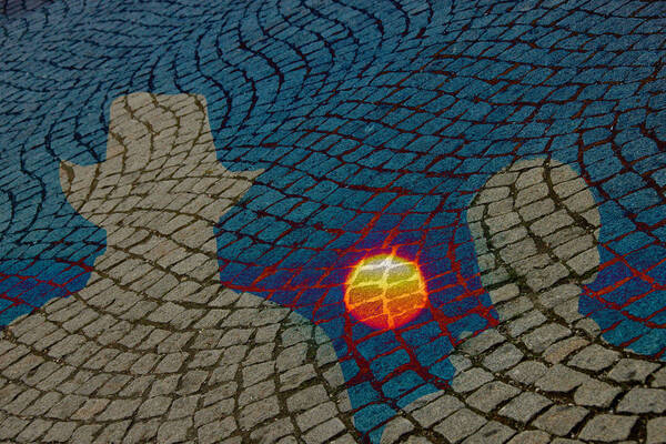 Die Silhouette zweier Menschen – eine Person davon trägt einen Hut – zeichnet sich hell auf einem Straßenpflaster von einem überlagerten Sonnenuntergang ab.