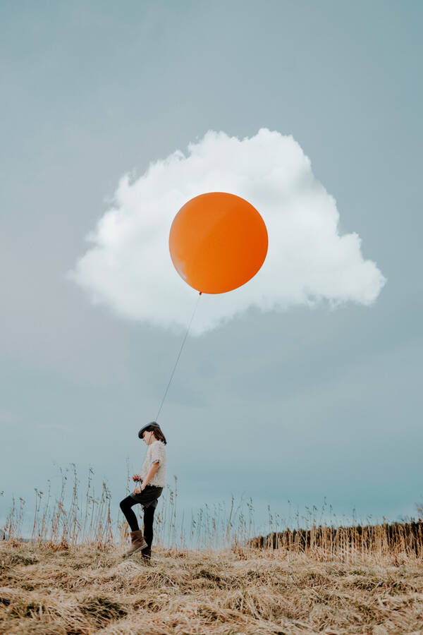 Eine Frau geht durch trockene Gräser und hält einen orange-farbenen Luftballon. Der Luftballon ist mitten in einer Wolke. Die Kombination aus Wolke und Luftballon erscheint wie ein Spiegelei.