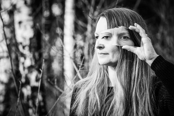 Eine Frau vor Bäumen blickt nach links aus dem Bild. In der linken Hand hält sie ein Fragment eines Bildes mit ihren Augen, die genau in die Kamera blicken. Schwarz-Weiß