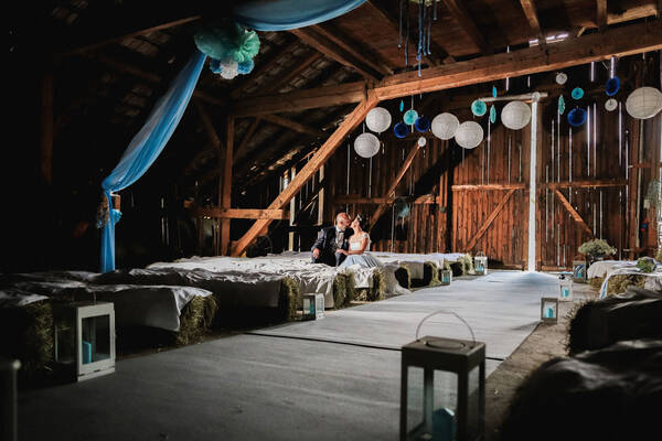 Ein Brautpaar sitzt auf mit weißen Tüchern bedeckten Strohballen in einer festlich dekorierten Scheune.