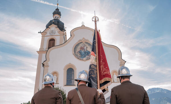 Drei Feuerwehrfunktionäre von hinten in Galauniform mit silberenen Helmen und mit einer Fahne vor einer Kirche.