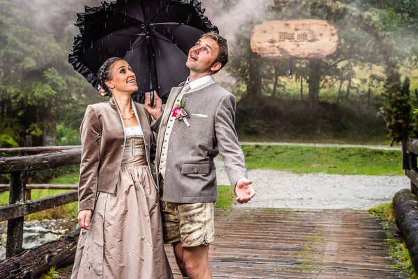Ein Paar in Tracht steht in nebliger Landschaft mit einem Regenschirm auf einer Brücke und blickt zum Himmel. Hinter Ihnen auf einem mit Grünzeug umrankten Bogen steht: 