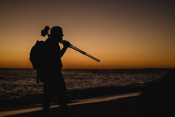 Silhouette eines Mannes mit Rucksack und einem Kamera-Stativ über der Schulter vor rot gefärbtem Himmel am Meer.