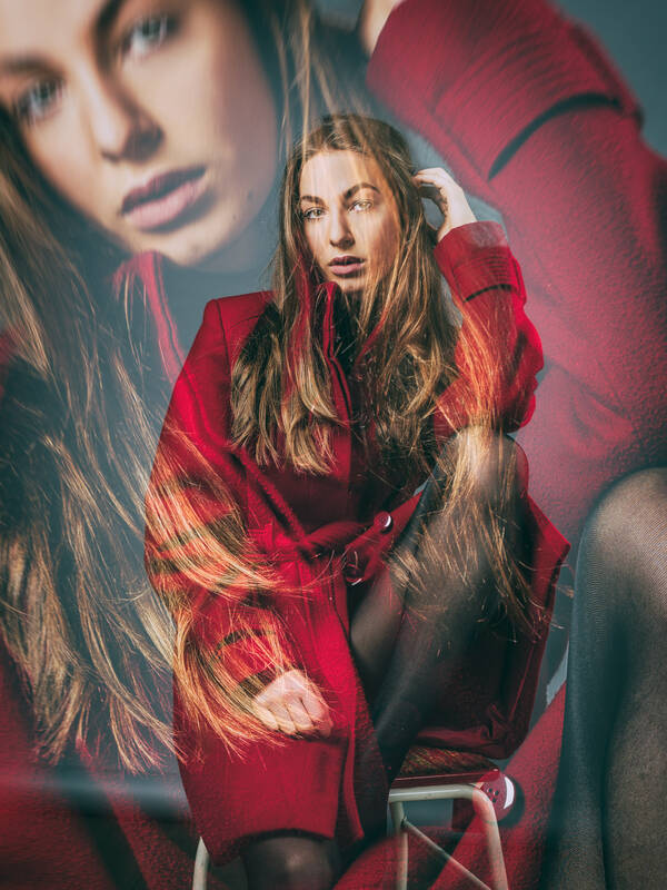 Eine junge Frau mit langen brünetten Haaren sitzt in einem roten Mantel und schwarzen Strümpfen auf einem Barhocker, das linke Bein angezogen und den linken Ellbogen darau abgestützt. Doppelbelichtung