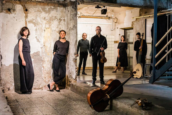 Ein sechsköpfiges Ensemble aus drei Frauen und drei Männern posiert in einem heruntergekommenen Gebäude. Zum Teil halten sie Instrumente wie Geigen oder Lauten, manche Instrumente wie Cello oder Bratsche liegen im Vordergrund.