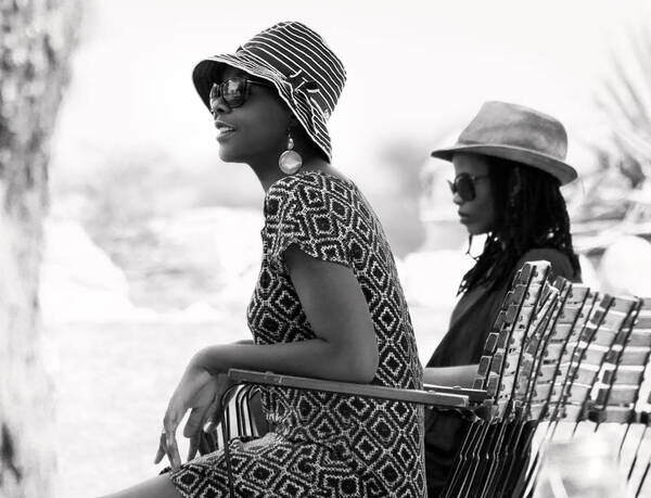 Zwei elegant, sommerlich gekleidete dunkelhäutige Frauen sitzen mit Sonnenbrille und Sonnenhot auf Grartensesseln. Schwarz-Weiß.