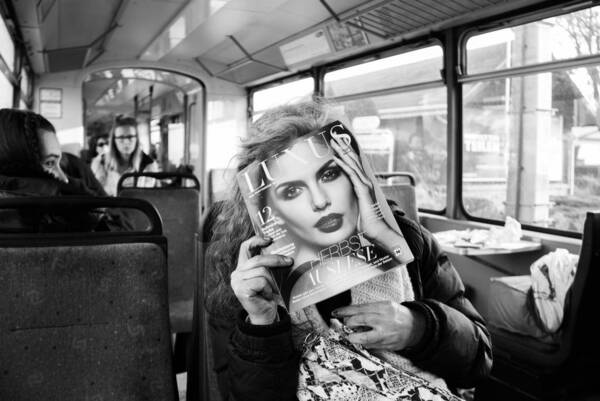 Eine alte Frau sitzt in der Straßenbahn und liest in der Zeitschrift Luxus. Das Portrait auf dem Titelblatt verdeckt ihr Gesicht.s.  Schwarz-Weiß.