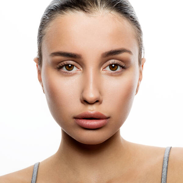 Closeup Portrait einer jungen Frau, die direkt in die Kamera schaut, die Haare streng nach hinten gekämmt. An den Schultern sieht man silbern glitzernde Trägerriemchen.