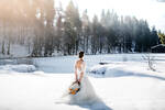 Eine Braut steht im Schnee am Rande eines zugefrorenen Sees. sie hält einen Blumenkranz mit beiden händen hinter ihrem Rücken.